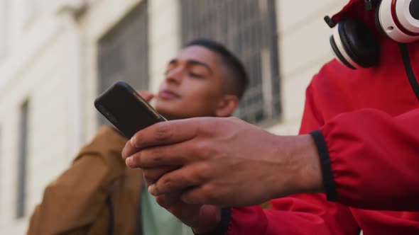 Two happy mixed race male friends sitting and using smartphone in the street