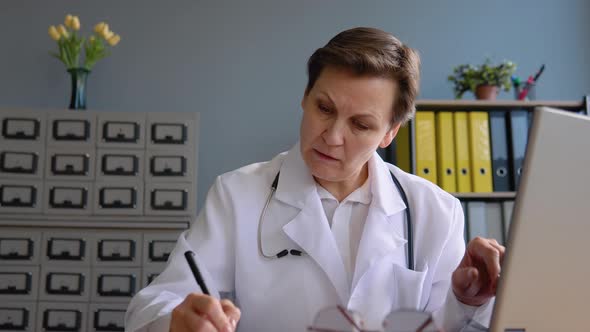 Senior Woman Doctor 50s Works with a Laptop and Writes Down the Patient's Medical History