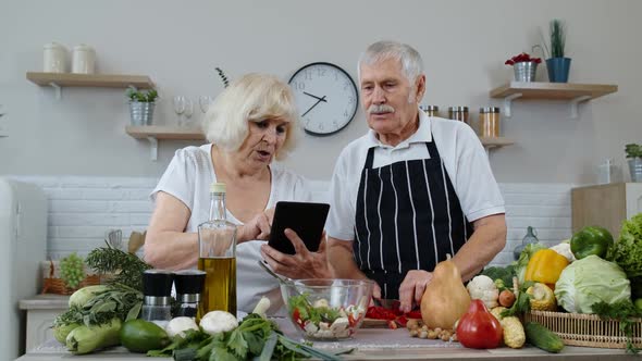 Vegan Senior Couple Cooking Salad with Raw Vegetables. Looking on Digital Tablet for Online Recipe