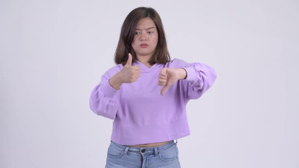 Young Confused Asian Woman Choosing Between Thumbs Up and Thumbs Down