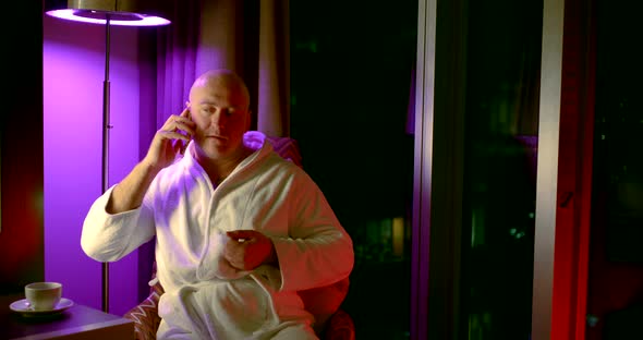 Bald Adult Man in White Robe Sits in a Chair in the Evening at Home and Speaks on the Phone