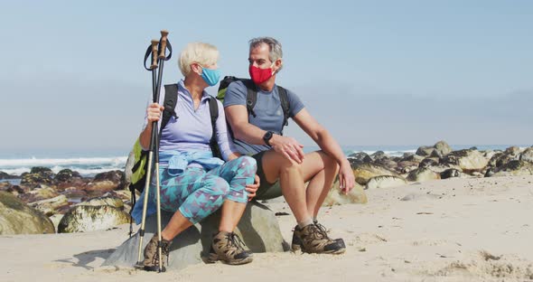 Senior hiker couple wearing face masks with backpacks and hiking poles talking sitting on rock