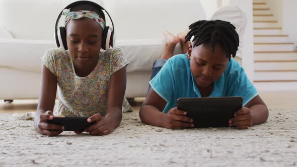 African american brother and sister using electronic devices lying on the floor at home