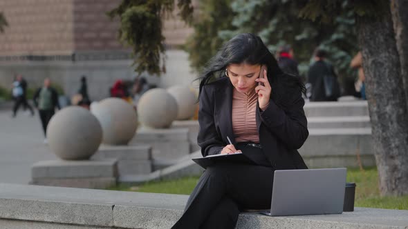 Young Multitasking Hispanic Businesswoman Freelancer Sitting Outdoors Talking on Mobile Phone