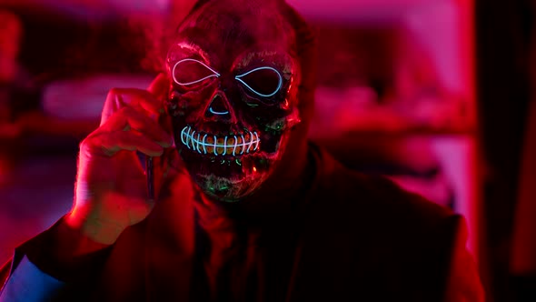 a Man in a Scary Glowing Death Mask Holds a Phone to Head Smokes and Nods in a Neon Flashing Light