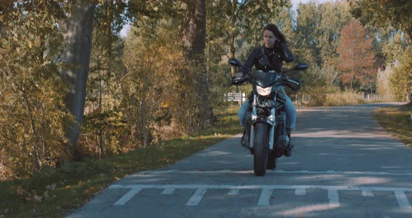 Pretty smiling European young woman driving a motorbike wearing leather jacket in forest with vibran