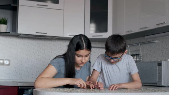 Cheerful Boy and Girl Will Put Together a Puzzle While Sitting at a Table at Home in the Kitchen