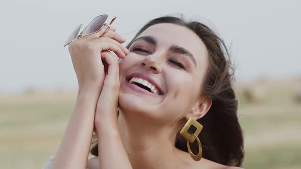 Portrait of Happy Woman Smiling Light at Windy Nature with Sunglasses in Hands