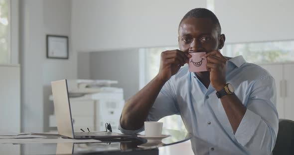 Medium Shot of African Businessman Holding Sticker with Drawn Smile By Mouth Looking at Camera