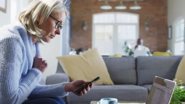 Worried senior caucasian woman in living room sitting on sofa, using smartphone and laptop