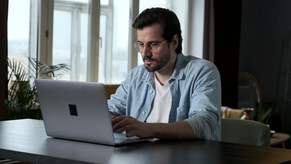 Smart Man in Glasses, Blue Shirt and White t-Shirt Working on a Computer laptop Online