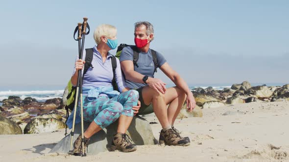 Senior hiker couple wearing face masks with backpacks and hiking poles talking sitting on rock