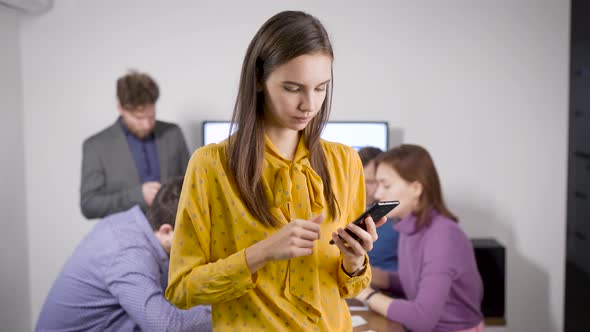 Young Woman Is Typing Message on Smartphone Standing in Office Room, Colleagues Are Sitting in
