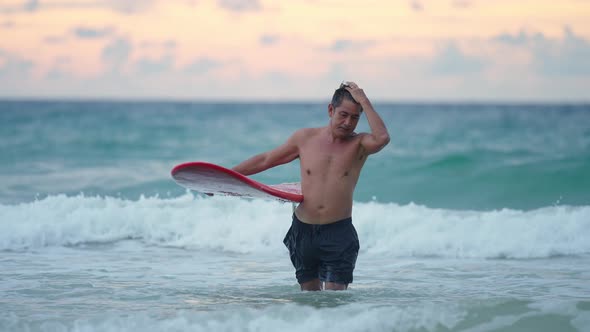 4K Asian senior man holding surfboard walking on the beach at summer sunset.