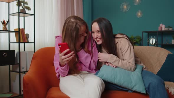 Cheerful Girls Friends Siblings Watching Photos on Smartphone on Sofa at Home Enjoy Social Media