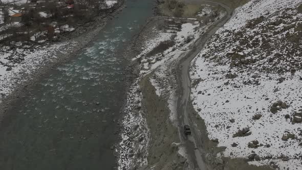 Aerial Over River In Hunza Valley Beside SUV Travelling Along Dirt Road On Snow Covered Hillside. Tr