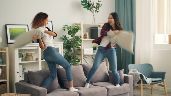 Playful Young Women African American and Asian Are Fighting with Pillows Standing on Sofa and