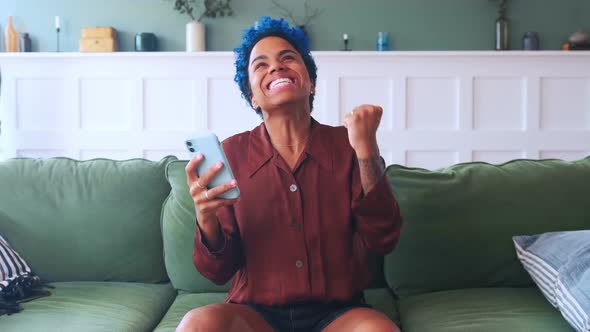 Young Overjoyed African American with Mobile Phone Sits on Sofa in Living Room