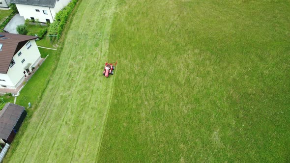 Aerial. Red lawnmower moving green meadows beside urbane settlement.