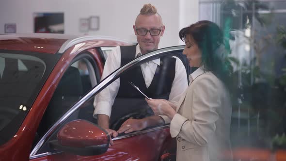 Serious Confident Caucasian Man Standing at Open Car in Dealership As Professional Woman Talking