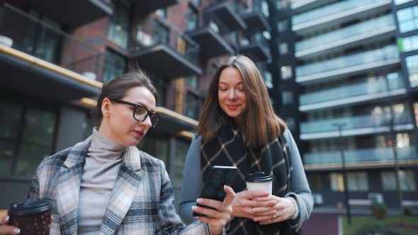 Two Happy Women Walking with Takeaway Coffee and Talking with Interest Among Themselves in the