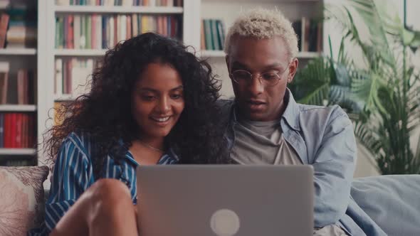 Happy Mixed Race Young Couple Using Laptop Looking at Screen Together on Sofa