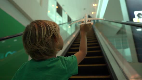 Slowmotion Shot of a Little Boy Moves Up the Escalator Holding White Toy Airplane in His Hand