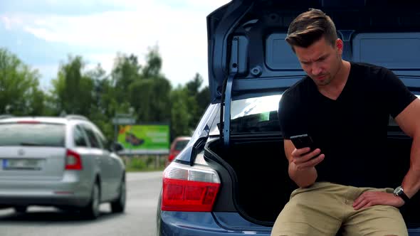 Young Handsome Man Sits in Car Trunk and Works (Typing) on Smartphone - Road 