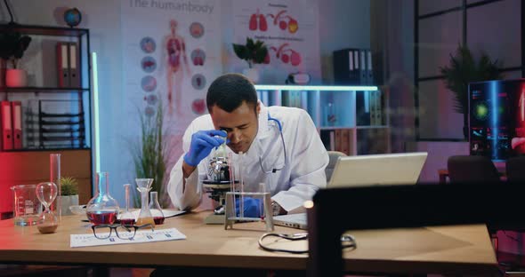 Male Chemist Viewing Samples Under Microscope and Noting Results of Experiment Into Report