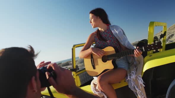 Caucasian man taking pictures of woman sitting in beach buggy by the sea playing guitar