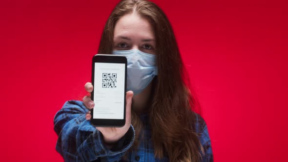 Woman Wearing Medical Mask Showing on Phone Vaccination Passport with Qr Code on Red Background