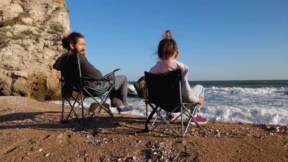 Father with Little Daughter Sits on Chairs on the Beach Rear View