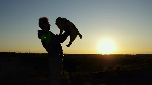 Man Tossing a Happy Pug Dog in the Air Hugs Over Sunset Sky