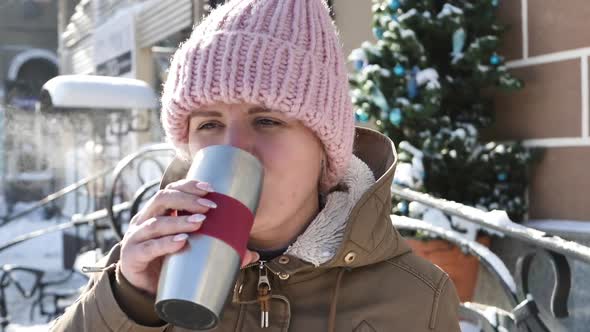 Young Beautiful Girl in a Warm Hat Drinks Tea From a Thermocup on the Street and Smiles