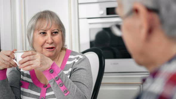 Smiling Aged Woman Speaking with Mature Husband Having Coffee Break at Kitchen Home