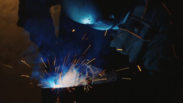 A Welder in a Protective Helmet and Clothes Welds Metal Sparks Illuminate It