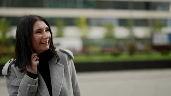 Brunette Woman in Grey Coat is Talking By Phone and Laughing Walking on Street