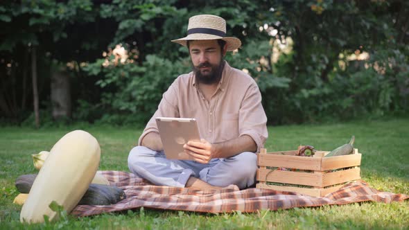 Smiling Positive Man Surfing Social Media on Tablet Sitting on Green Meadow Outdoors with Harvest