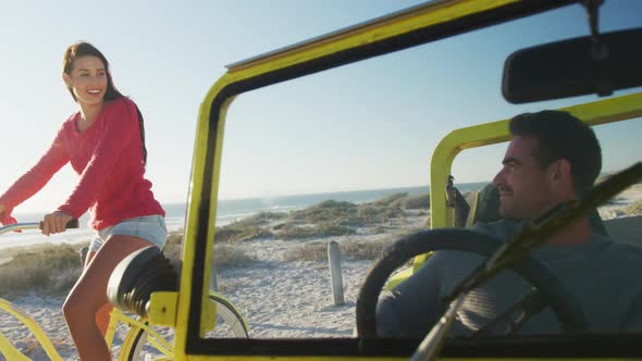 Happy caucasian woman riding a bike next to man in beach buggy by the sea