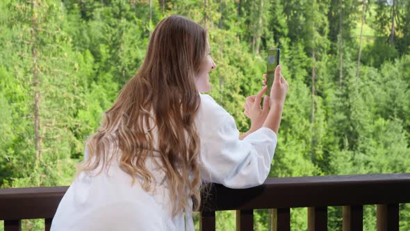 Beautiful Young Woman in Bathrobe Standing on Balcony and Making Image of Mountains and Forest on