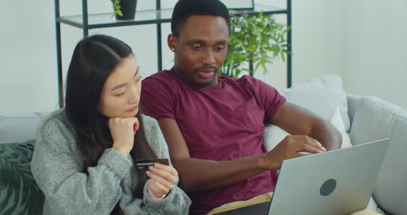Happy Young Mixed Ethnicity Couple Spend Time at Modern Home Using Laptop Buying Via Credit Card