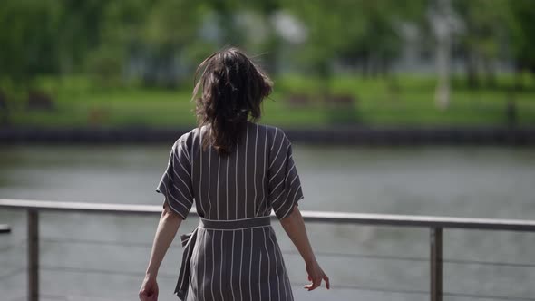 a Brunette in a Striped Dress Approaches the Railing of a Bridge Over the River on a Windy Sunny Day