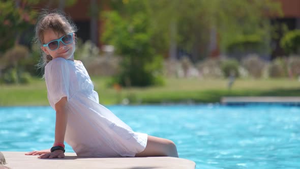 Young Joyful Child Girl in White Dress Resting on Swimming Pool Side with Clear Blue Water on Sunny