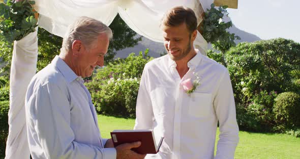 Smiling caucasian senior male wedding officiant holding book and groom standing in outdoor altar