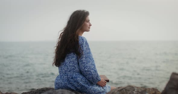 A Young Woman Sits on a Large Stone on the Seashore and Looks Into Sea Distance