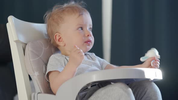 Cute Kid with Food Nibbler Baby Fruit Pacifier Sitting in Booster Seat Fixed on Top of Dining Chair