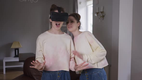 Happy Absorbed Young Woman in VR Headset Enjoying Augmented Reality As Curios Twin Sister Asking to