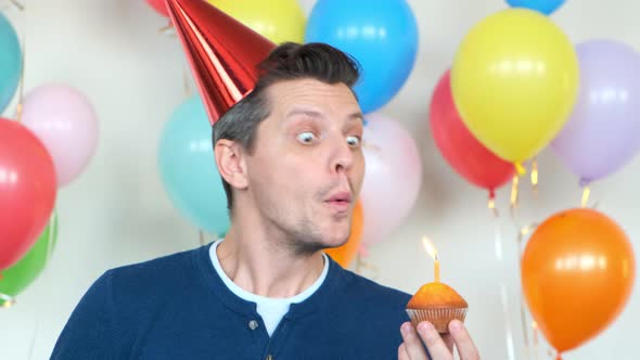 Funny Man in Red Party Cone Blows Burning Candle on Cupcake