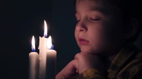 Little Cute Boy Reads a Prayer in the Dark with a Candle