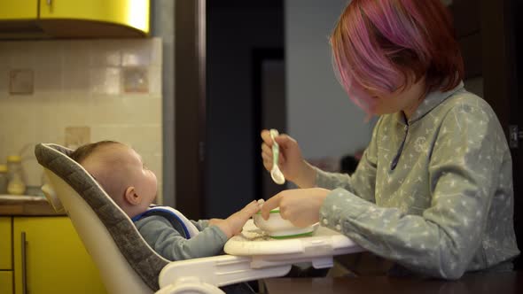 Mother is Giving Porridge to Her Son Sitting in the Highchair in the Kitchen During the Breakfast